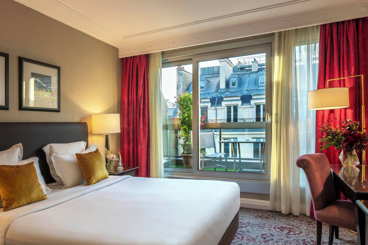 A hotel room with a large bed, two bedside tables, a chair, a window with curtains, and a view of buildings outside.