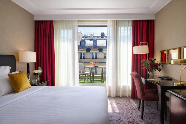 A hotel room with a bed, a desk, a chair, a lamp, and a view of the outside balcony.