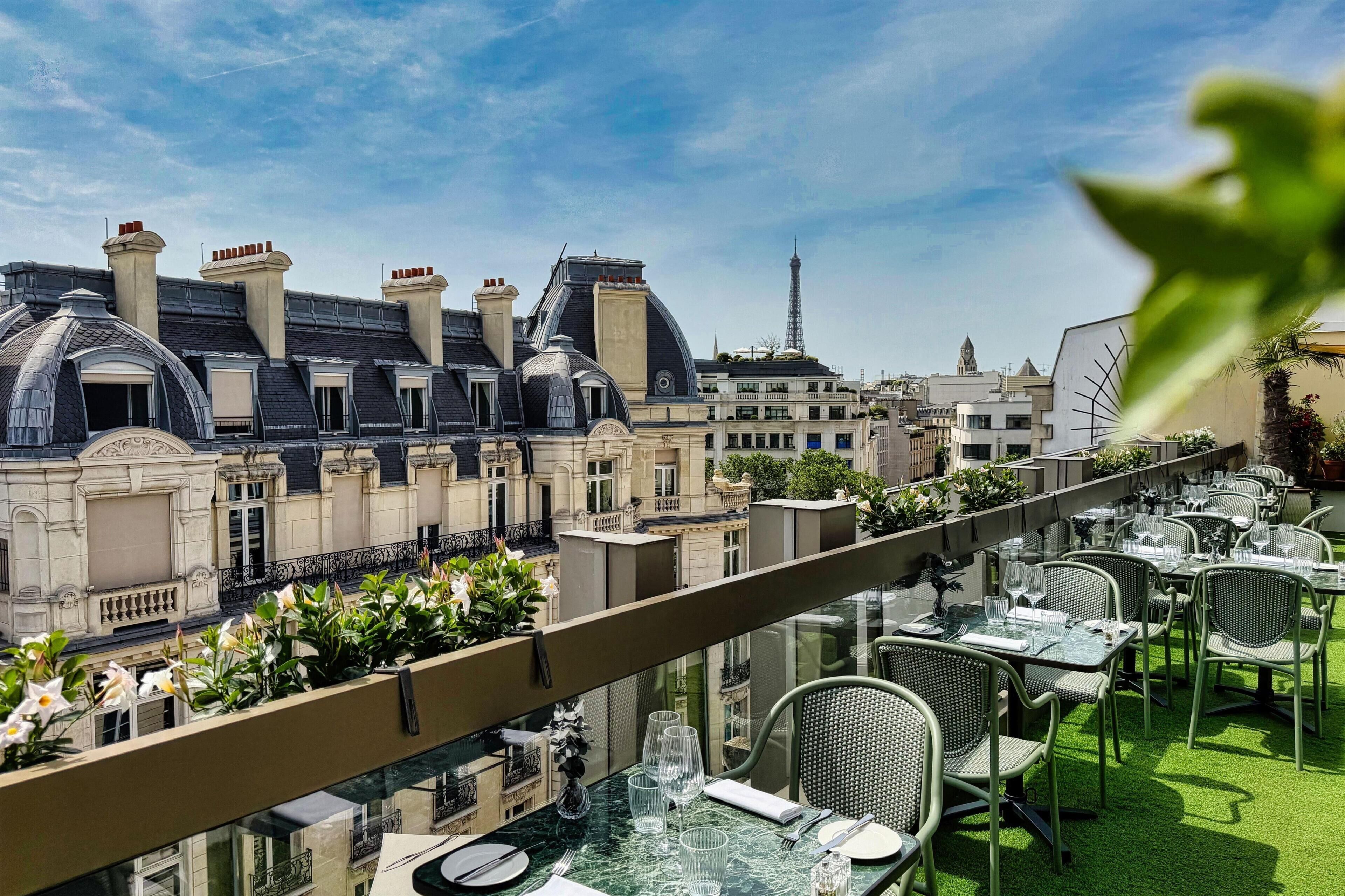 A rooftop terrace with tables and chairs, overlooking a cityscape with historic buildings and the Eiffel Tower in the distance.