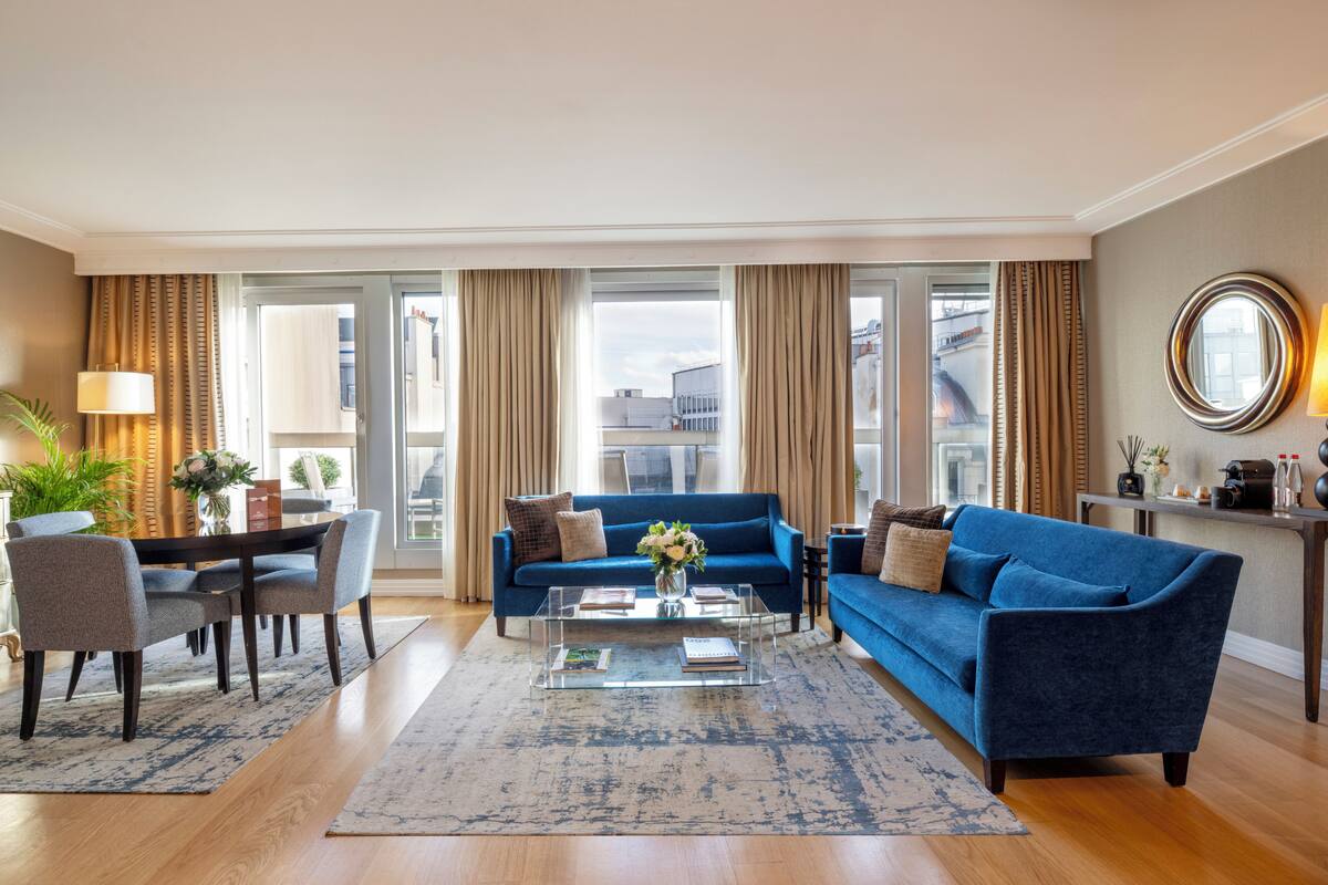 A modern living room with a blue sofa, a glass coffee table, and a dining area with a table and chairs.