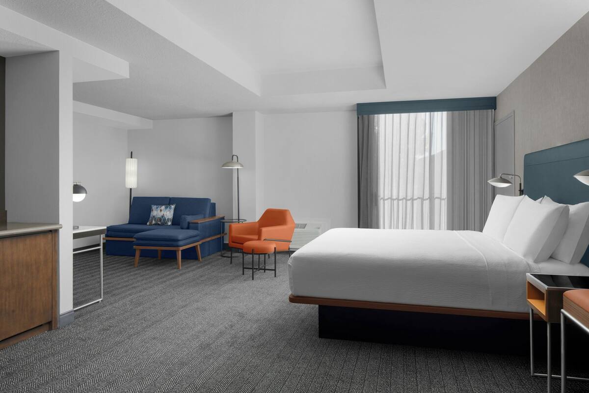 A modern hotel room with a large bed, a desk, a blue sofa, an orange chair, and a large window with curtains.