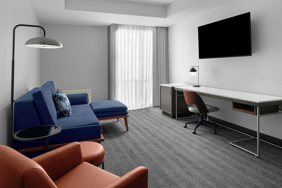 A modern hotel room with a blue sofa, orange armchair, a desk with a chair, a flat-screen TV, and a large window with sheer curtains.