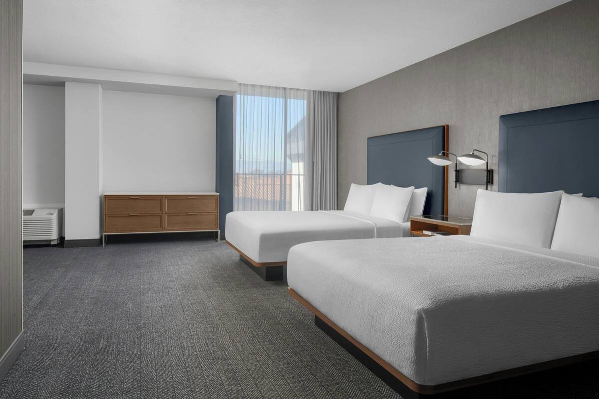 A hotel room with two beds, a wooden dresser, a desk with a lamp, and a large window with curtains.