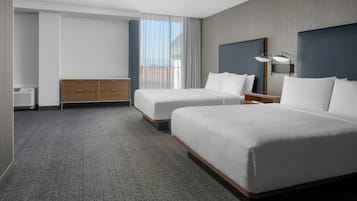 A hotel room with two beds, a wooden dresser, a desk with a lamp, and a large window with curtains.