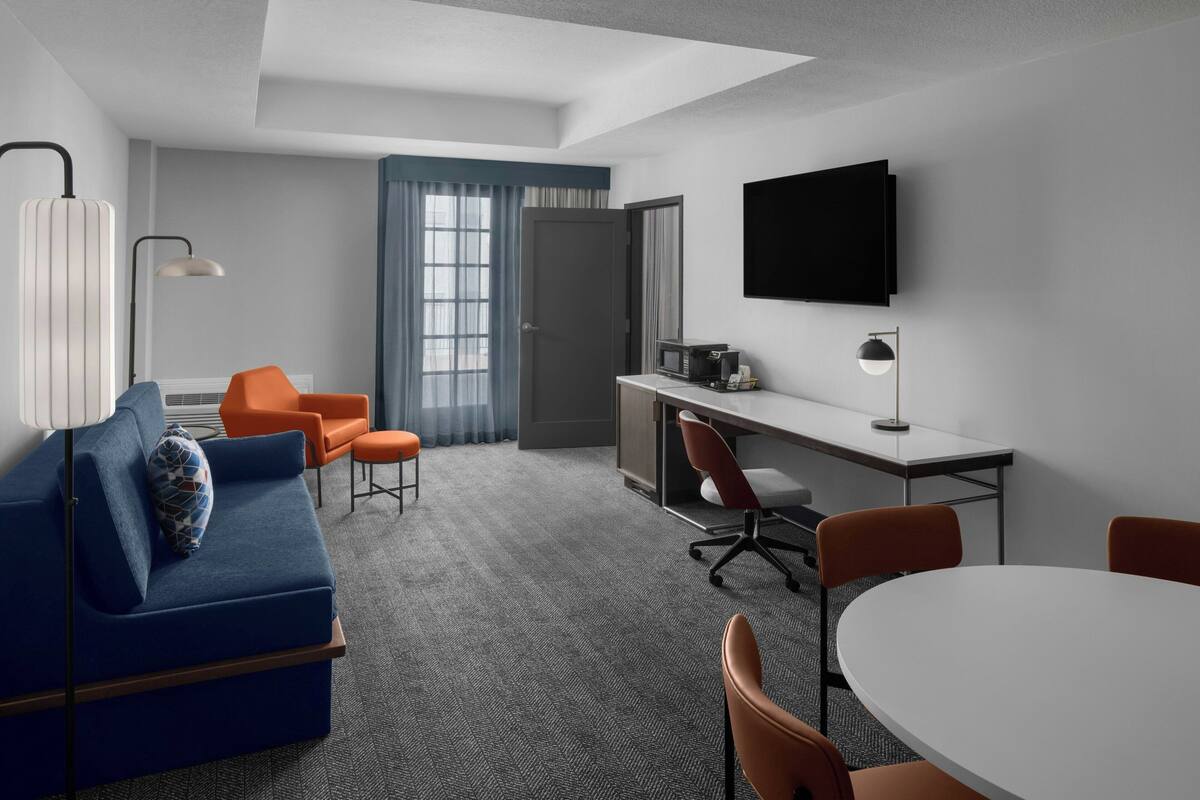 A modern hotel room with a blue sofa, orange chairs, a white table, and a TV mounted on the wall.
