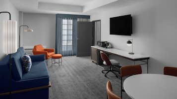 A modern hotel room with a blue sofa, orange chairs, a white table, and a TV mounted on the wall.