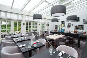 Un restaurante moderno con techos altos y amplios ventanales, que ofrece vistas al exterior. Las mesas están dispuestas con copas y cubiertos, y hay jarrones decorativos sobre ellas.