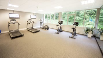 A gym with treadmills, elliptical machines, and stationary bikes, large windows with a view of greenery, and a flat-screen TV on the wall.