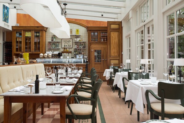A restaurant with multiple dining tables and chairs, a bar area with bottles and glasses, and a view through large windows. - Hotel De Bilderberg (Oosterbeek)