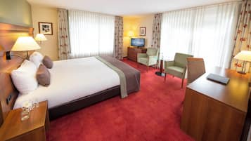 Een hotelkamer met een groot bed, twee stoelen, een bureau, een televisie en een kleine tafel met een bloevas.