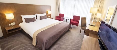 A hotel room with a large bed, two bedside tables with lamps, a TV, a desk with a chair, and a window with curtains.