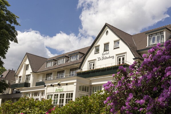 Un hotel con fachada blanca y numerosas ventanas, incluyendo un balcón con flores rojas.