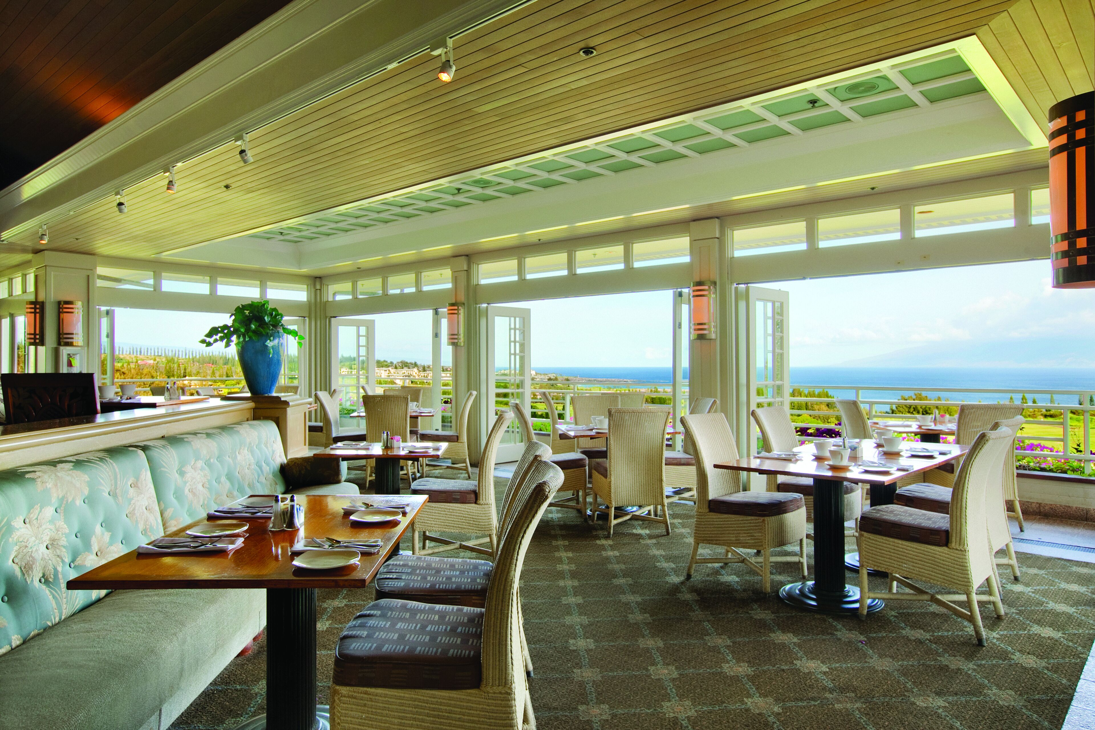 A spacious outdoor dining area with multiple tables and chairs, a large potted plant, and a view of the ocean.