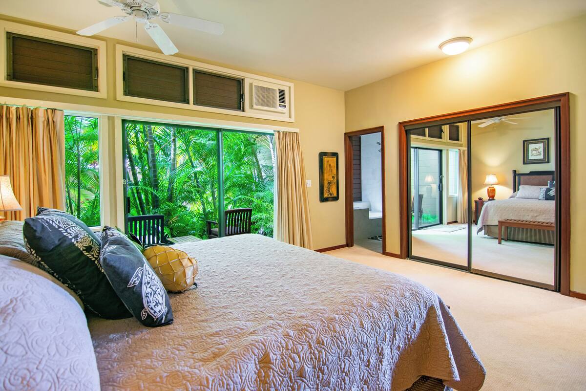 Ein Schlafzimmer mit einem großen Bett, einem Deckenventilator und Blick auf üppiges Grün durch das Fenster.
