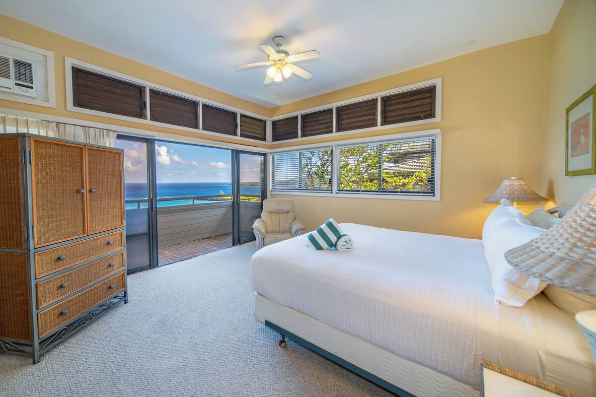 Ein Schlafzimmer mit einem großen Bett, einem Schminktisch, einem Deckenventilator und Blick aufs Meer durch die Fenster.