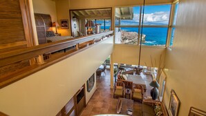 A spacious living area with a high ceiling, a balcony overlooking the ocean, and a view of rocky terrain.