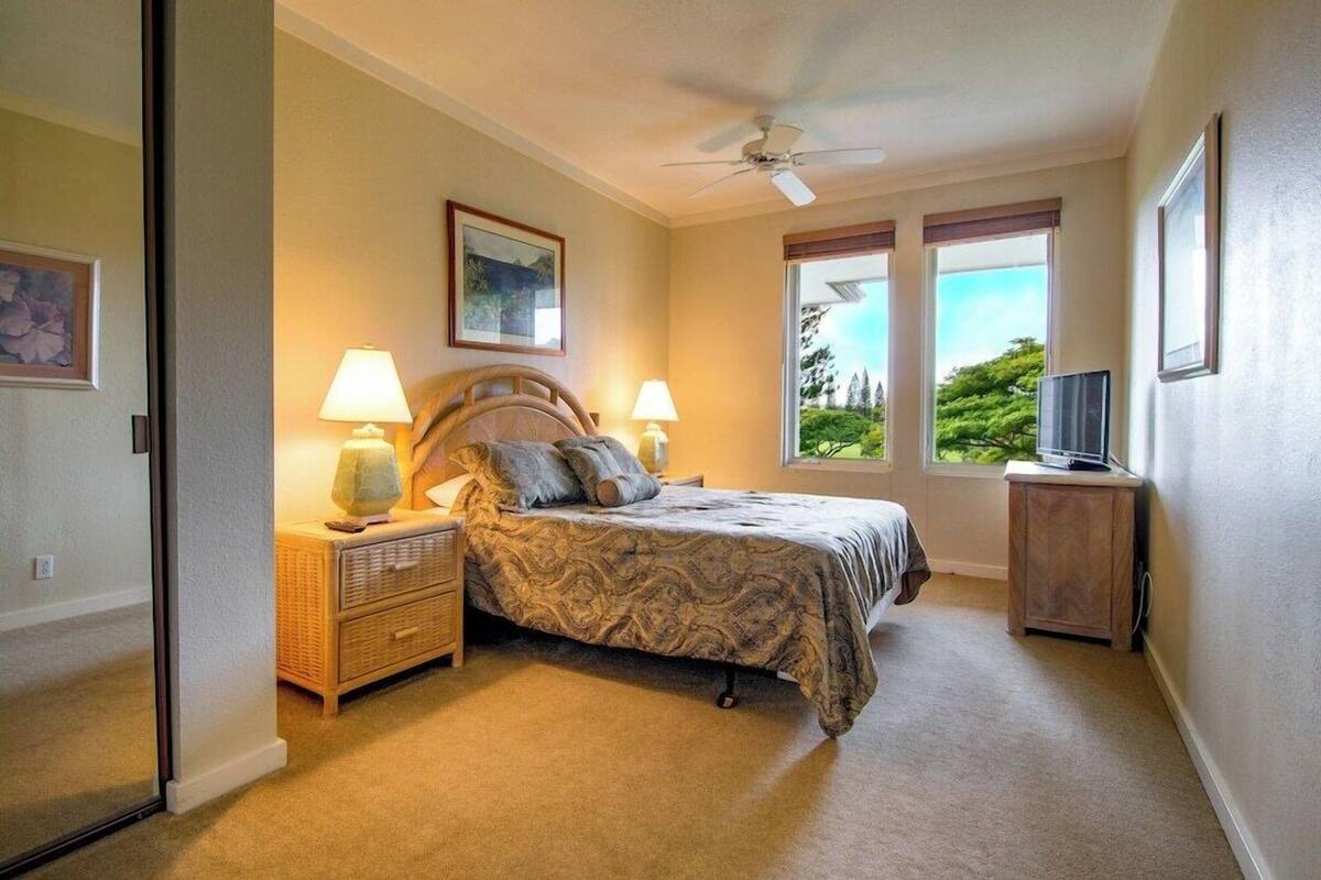 Ein Schlafzimmer mit einem Bett, zwei Nachttischlampen, einem Deckenventilator, einem Fernsehschrank und einem Fenster mit Blick auf Bäume.