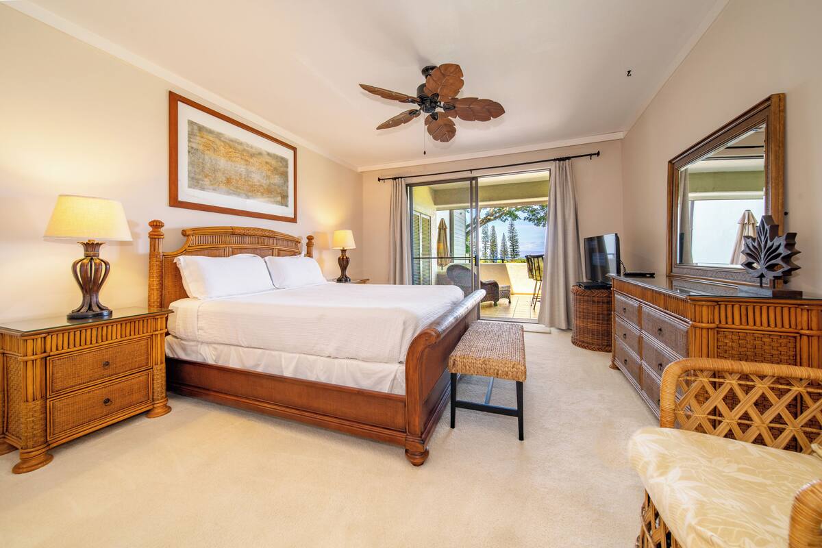 A bedroom with a wooden bed, two bedside tables with lamps, a ceiling fan, a large mirror, and a view of the outdoors through a sliding door.