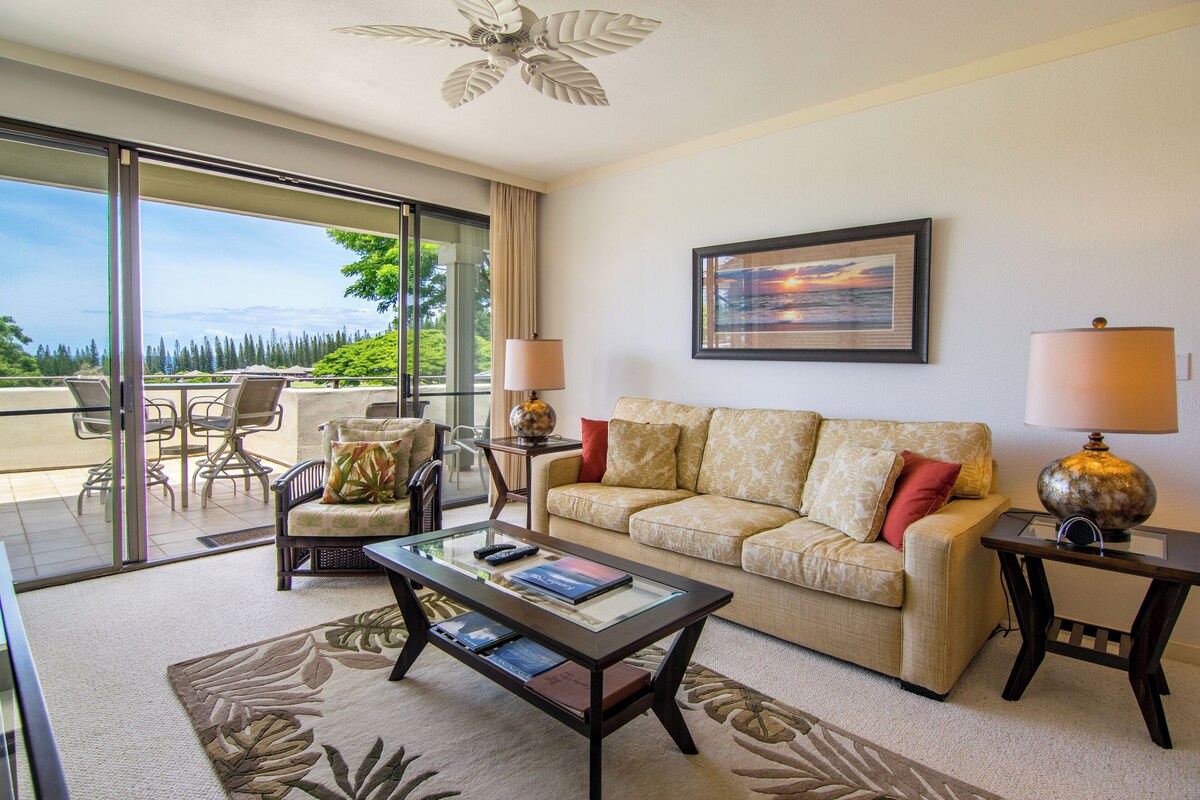 A spacious living room with a glass coffee table, a beige sofa with red cushions, a framed painting on the wall, and a view of the outdoors through large sliding doors.