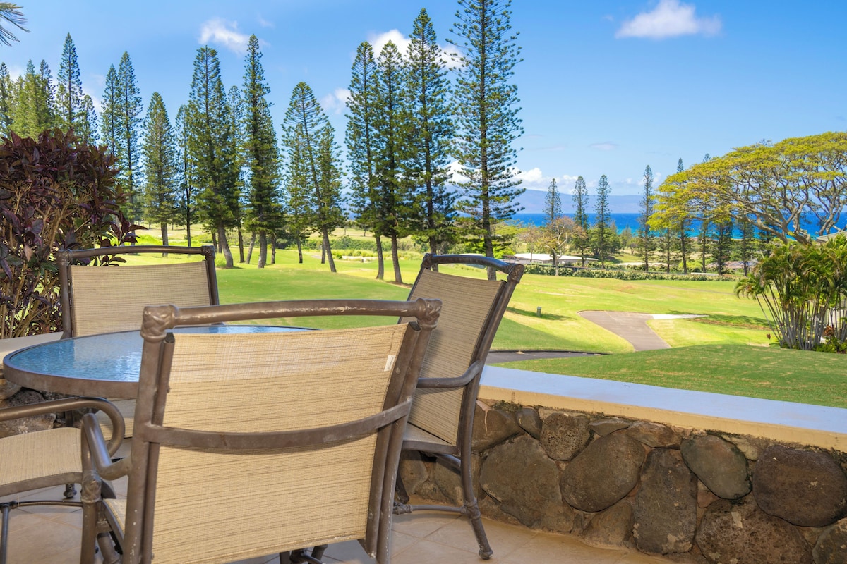 A patio with a round table and chairs, overlooking a golf course with a view of the ocean.