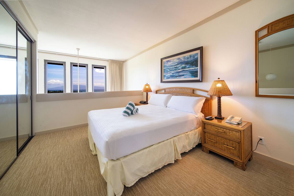 Ein Hotelzimmer mit einem großen Bett, Nachttischlampen, einem Nachttisch, einem Gemälde und Blick durch das Fenster.