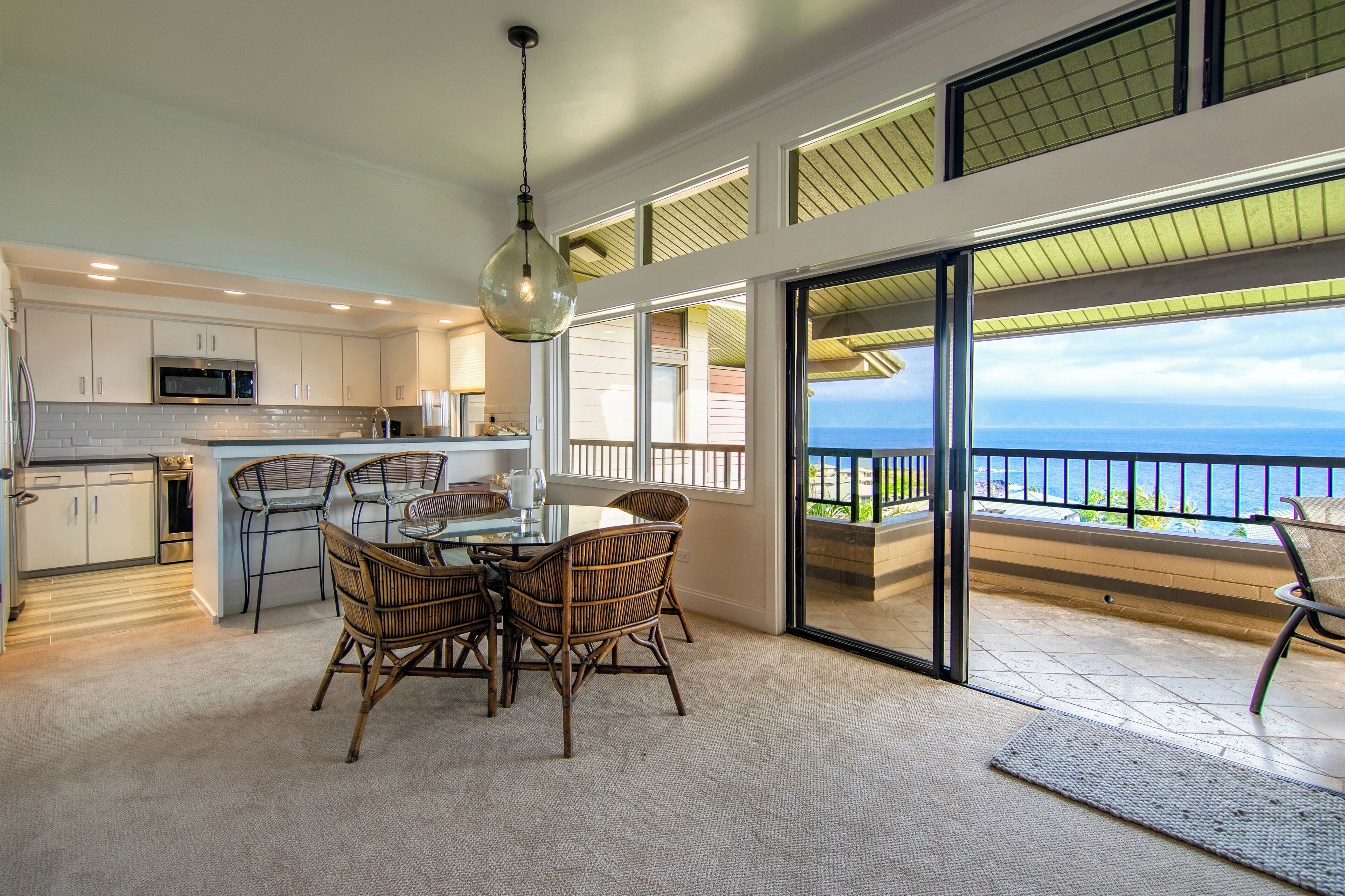 A modern kitchen with white cabinets and a large island, a dining area with wicker chairs, and a balcony with a view of the ocean.