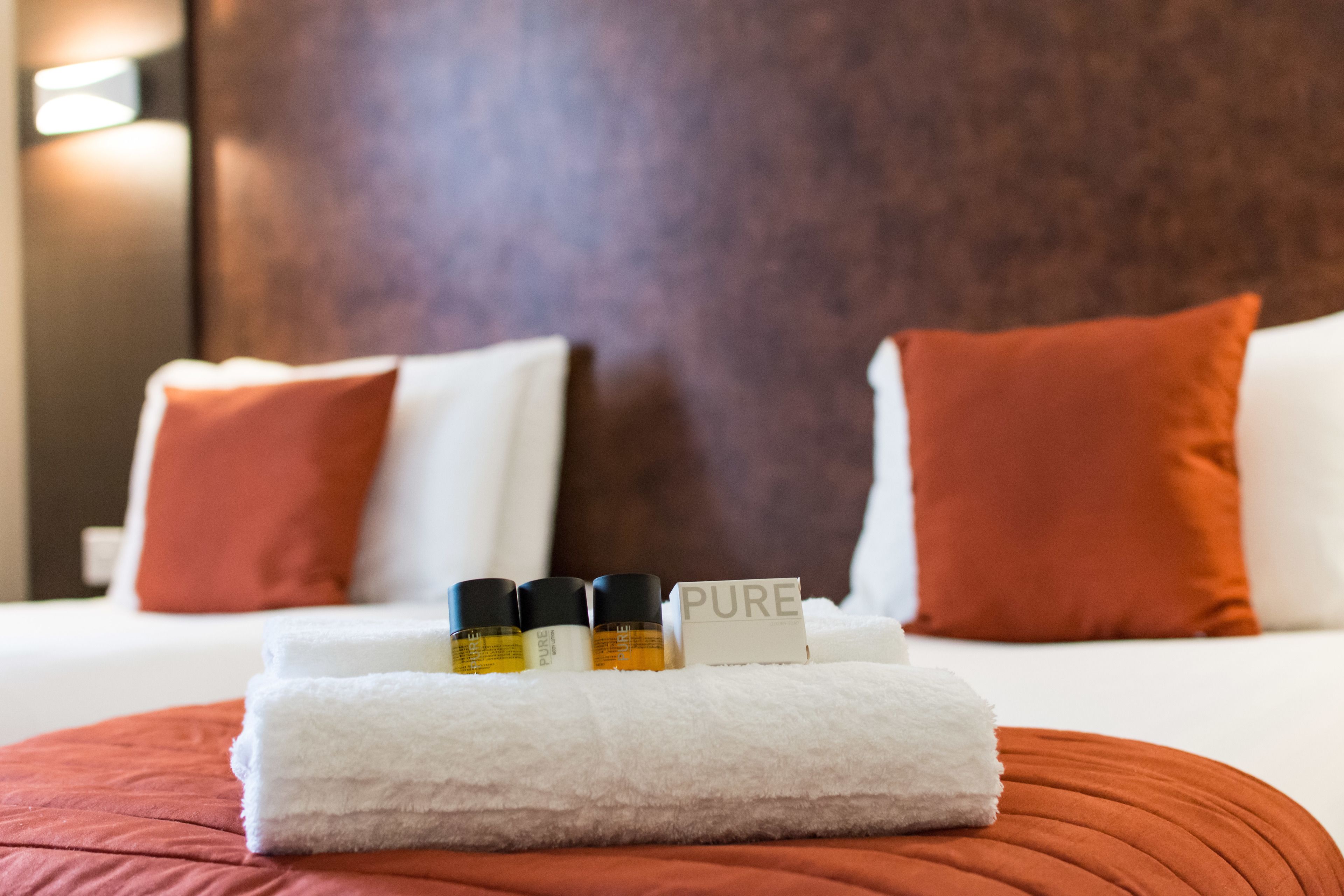 Una cama bien tendida con almohadas blancas y rojas, una toalla blanca y tres botellas con la etiqueta 'PURE' sobre un cojín rojo.