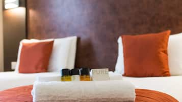 Una cama bien tendida con almohadas blancas y rojas, una toalla blanca y tres botellas con la etiqueta 'PURE' sobre un cojín rojo.