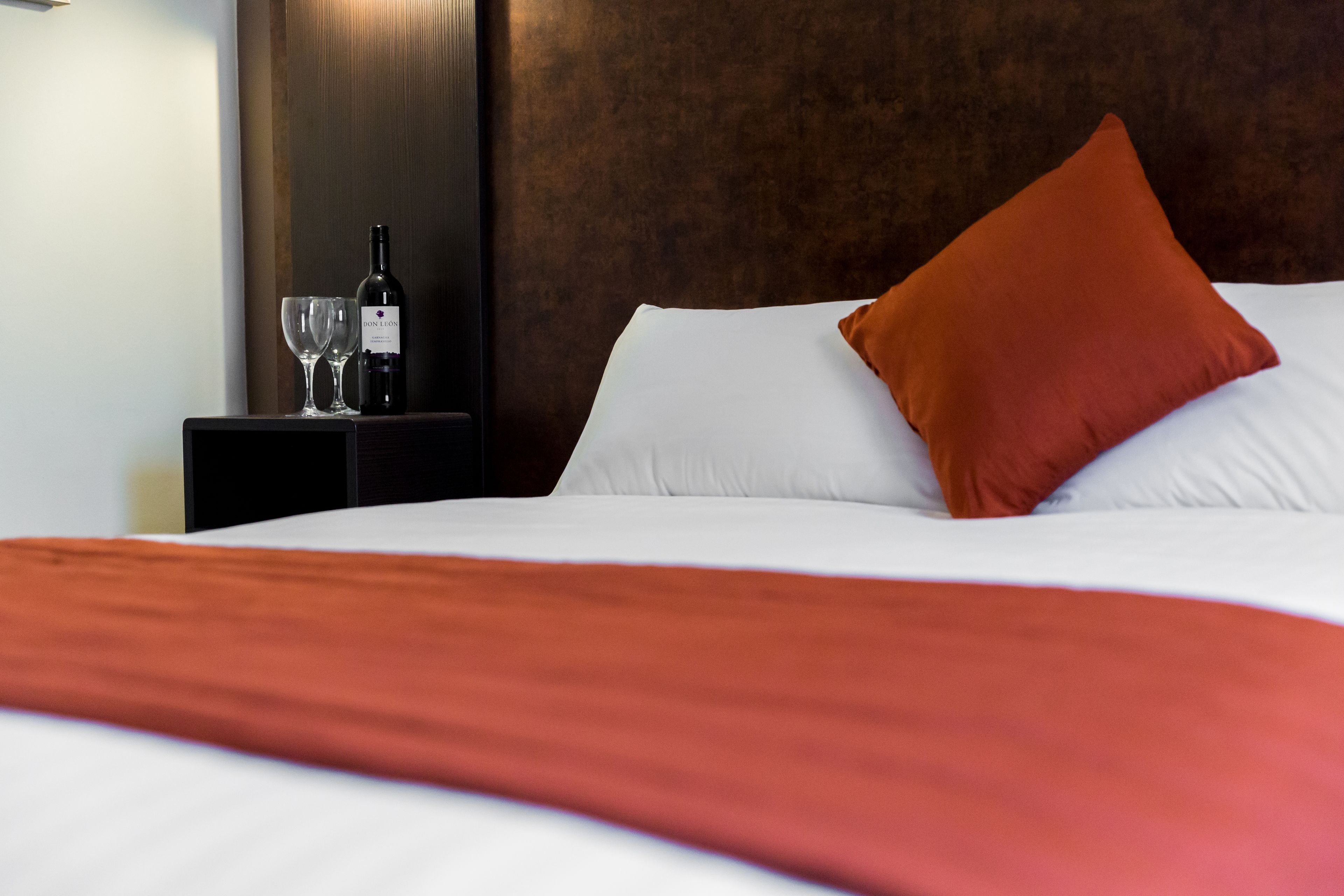 A neatly made bed with white and orange bedding, a bottle of wine, and two glasses on a small table beside it.