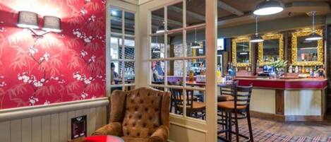 Un coin bar avec un papier peint à motifs floraux rouges, un fauteuil en cuir capitonné et un comptoir de bar avec des tabourets.