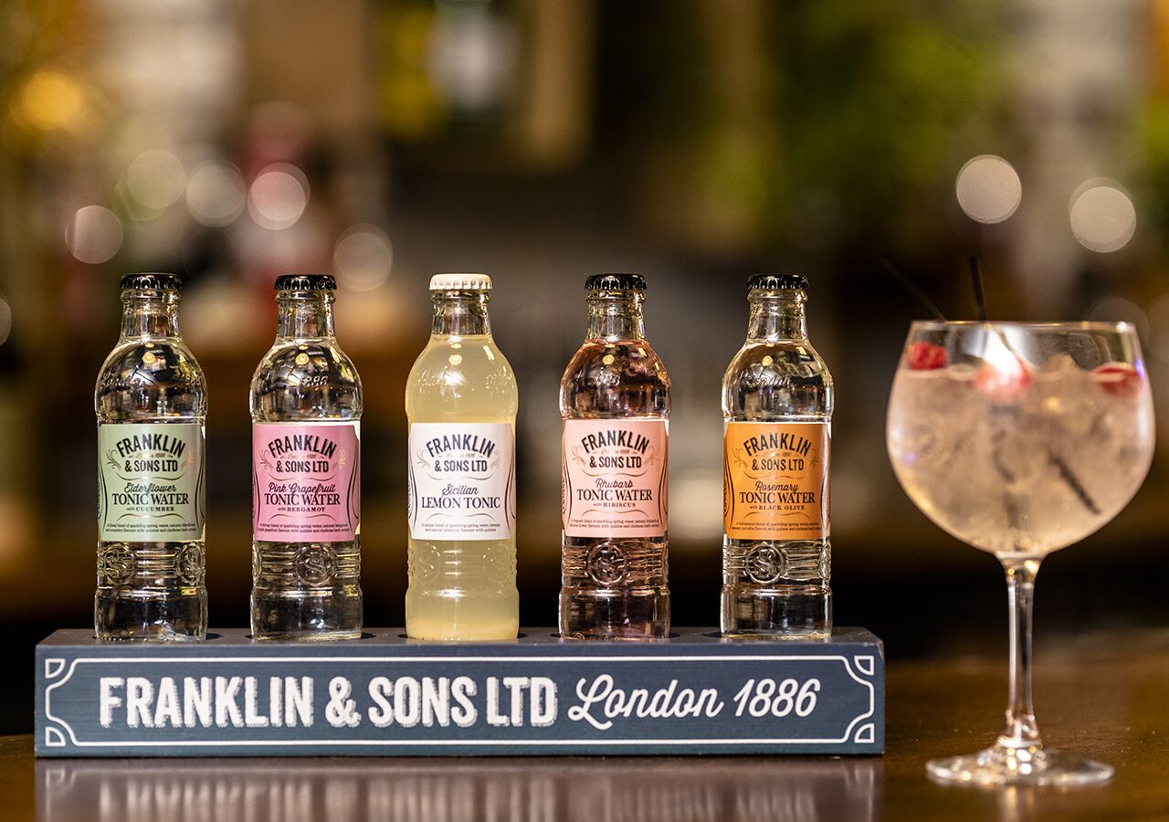 A display of five bottles of tonic water from Franklin & Sons Ltd, London 1886, with a glass of a mixed drink beside them.