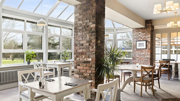 A spacious dining area with white wooden tables and chairs, a brick column, and large windows providing natural light. - Dragonfly Hotel King's Lynn (King's Lynn)