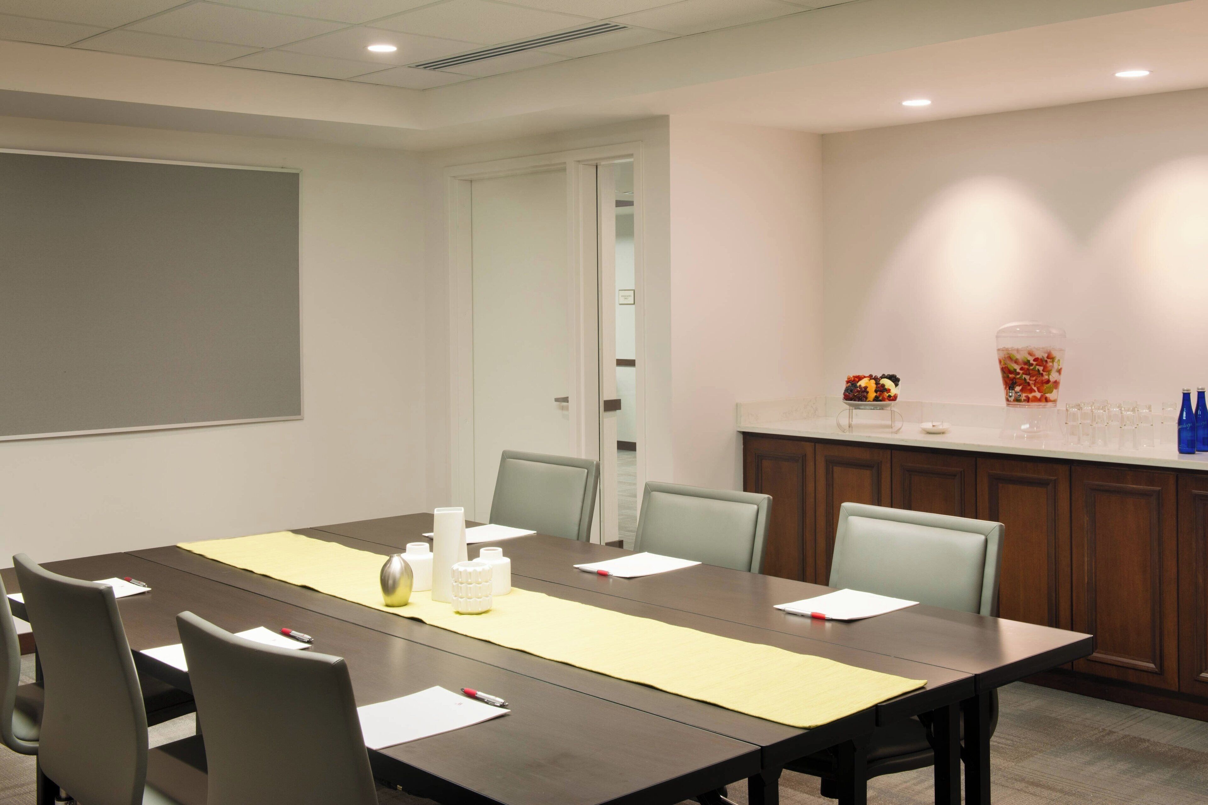 A conference room with a long table, chairs, a projector screen, and a small kitchenette area with a beverage dispenser and glasses.