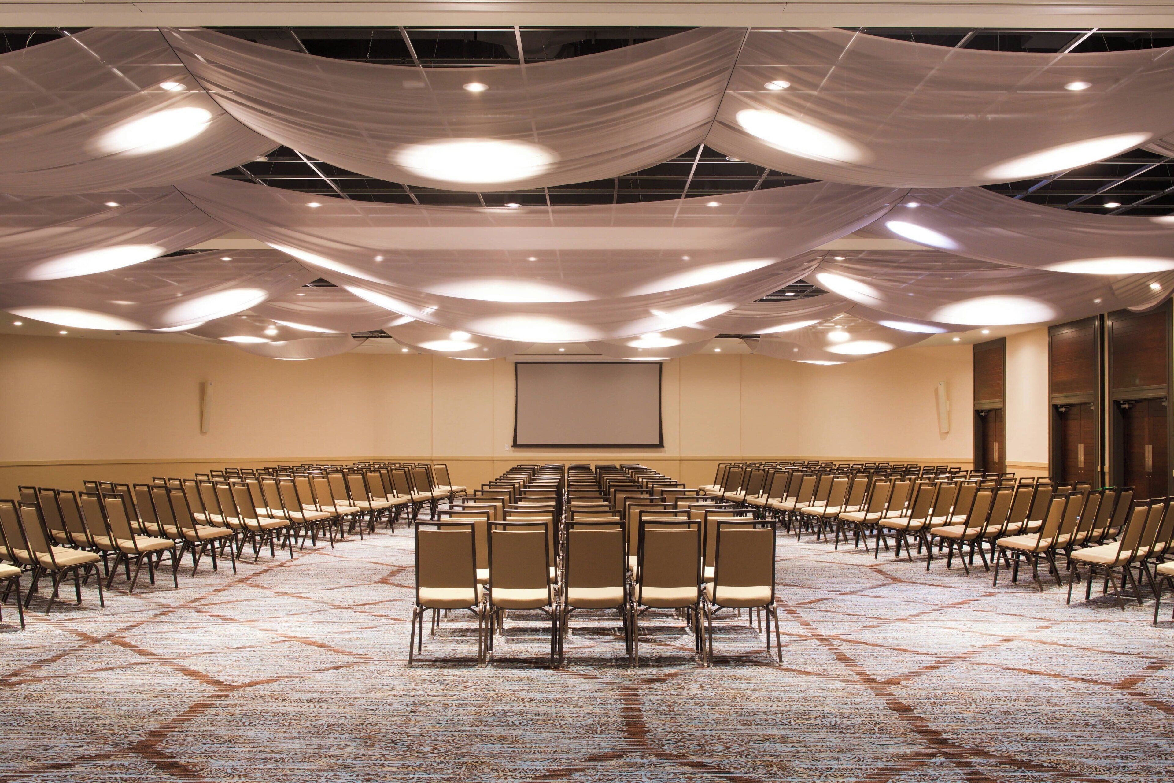 A conference room with rows of chairs facing a projection screen, a carpeted floor, and draped fabric on the ceiling.