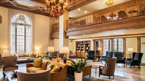 Un hall d’entrée spacieux avec un plafond haut, des balustrades décorées et un lustre. On y trouve plusieurs coins salon avec des canapés et des fauteuils, ainsi qu’une grande bibliothèque remplie de livres.