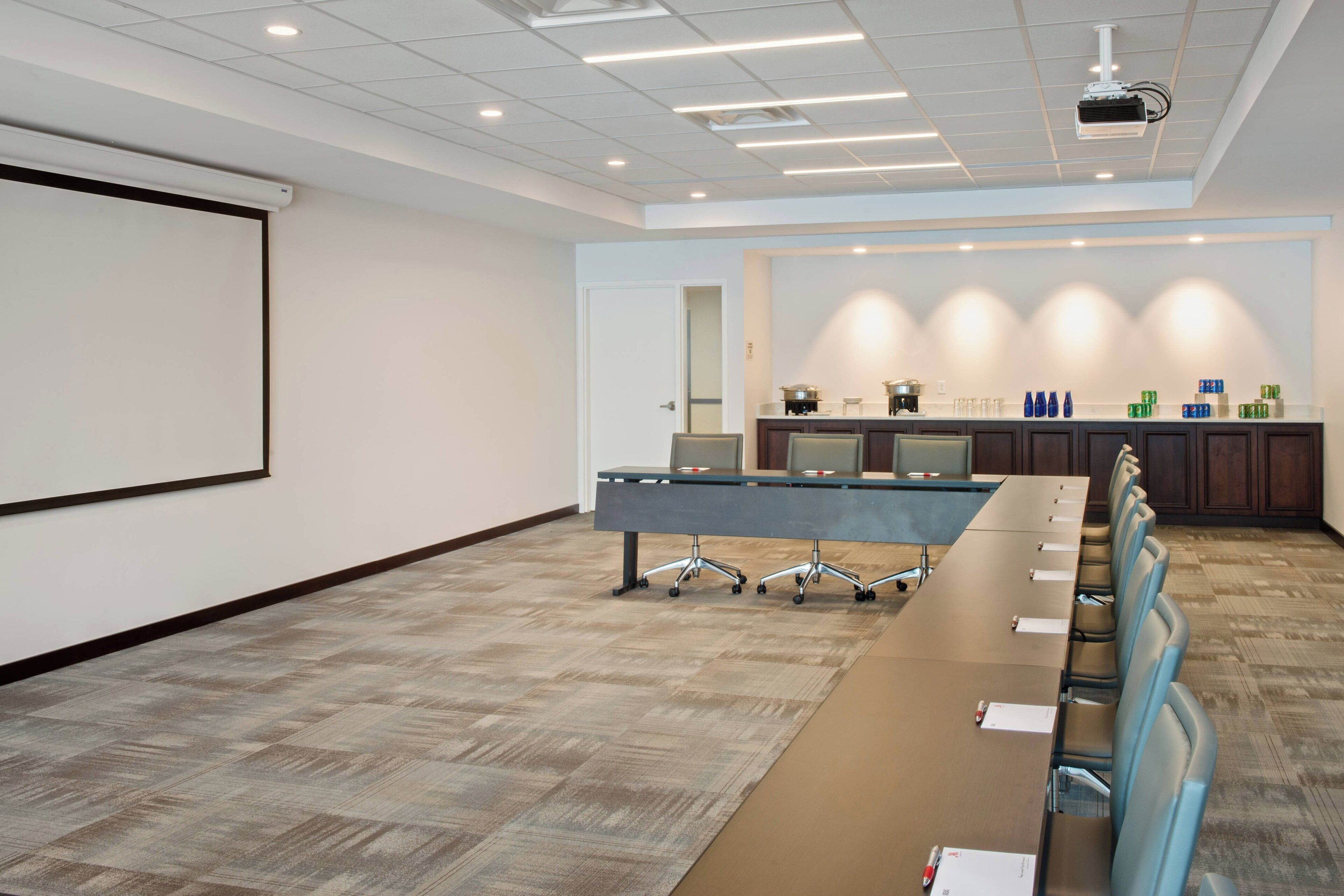 A conference room with a long table, chairs, a projector screen, and a kitchen area in the background.