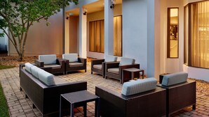 An outdoor seating area with wicker furniture and cushions, a paved patio, and a building with large windows and a tree. - Courtyard by Marriott - Atlanta Executive Park/Emory (Atlanta)