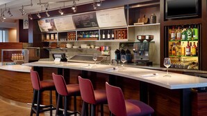 A bar area with a long counter, red bar stools, a coffee machine, and shelves with bottles and glasses. - Courtyard by Marriott - Atlanta Executive Park/Emory (Atlanta)
