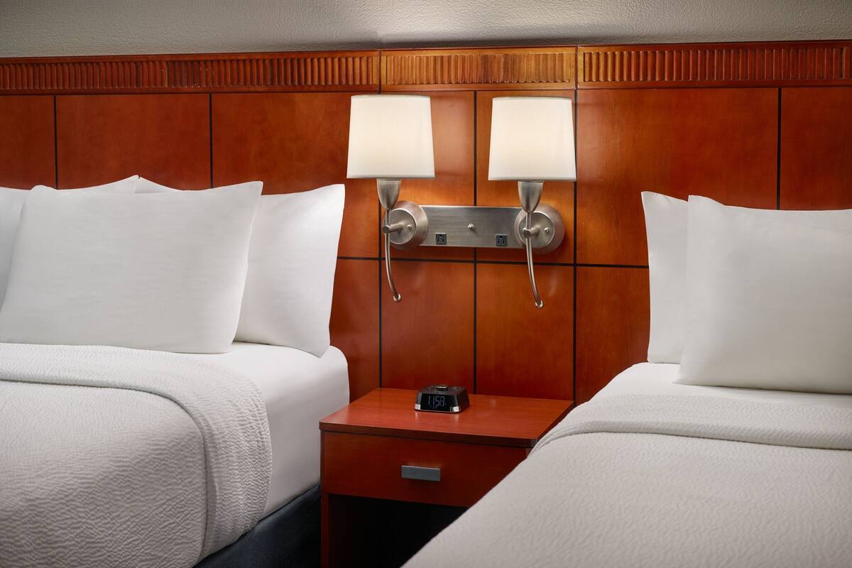 Habitación de hotel con dos camas, cabecera de madera, lámparas de noche, mesita de noche con un reloj y un cajón.