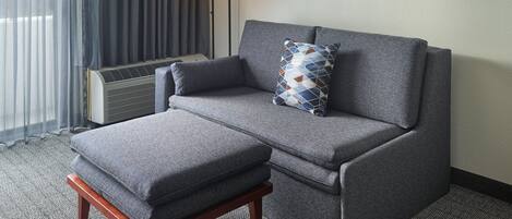 A grey sofa with a matching ottoman and a patterned pillow, a floor lamp, and a radiator in a hotel room.