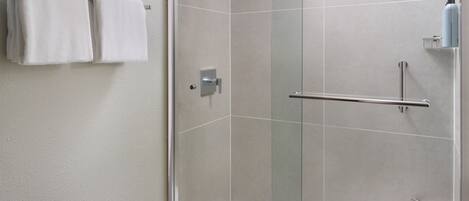 A glass-enclosed shower with a handrail, a towel rack with two white towels, and a small shelf with a blue bottle.