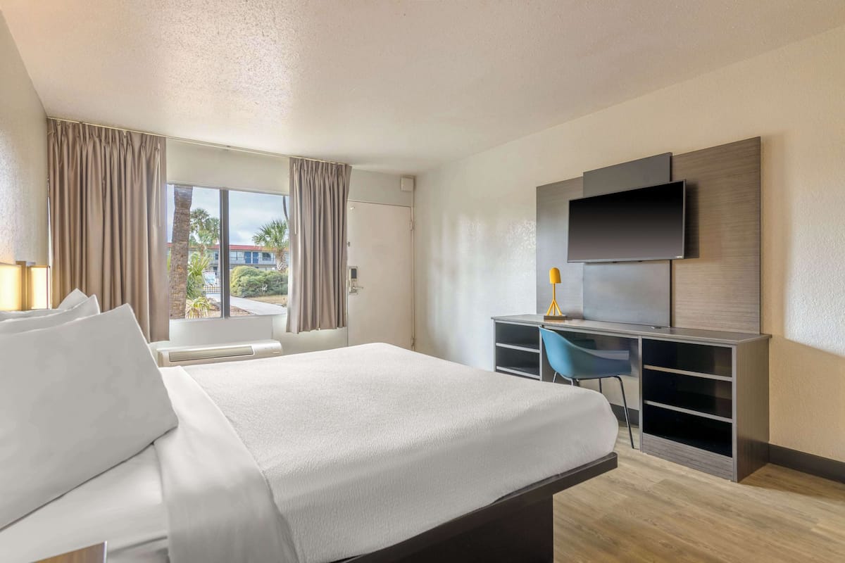 A hotel room with a bed, a desk with a chair, a TV mounted on the wall, and a window with curtains.