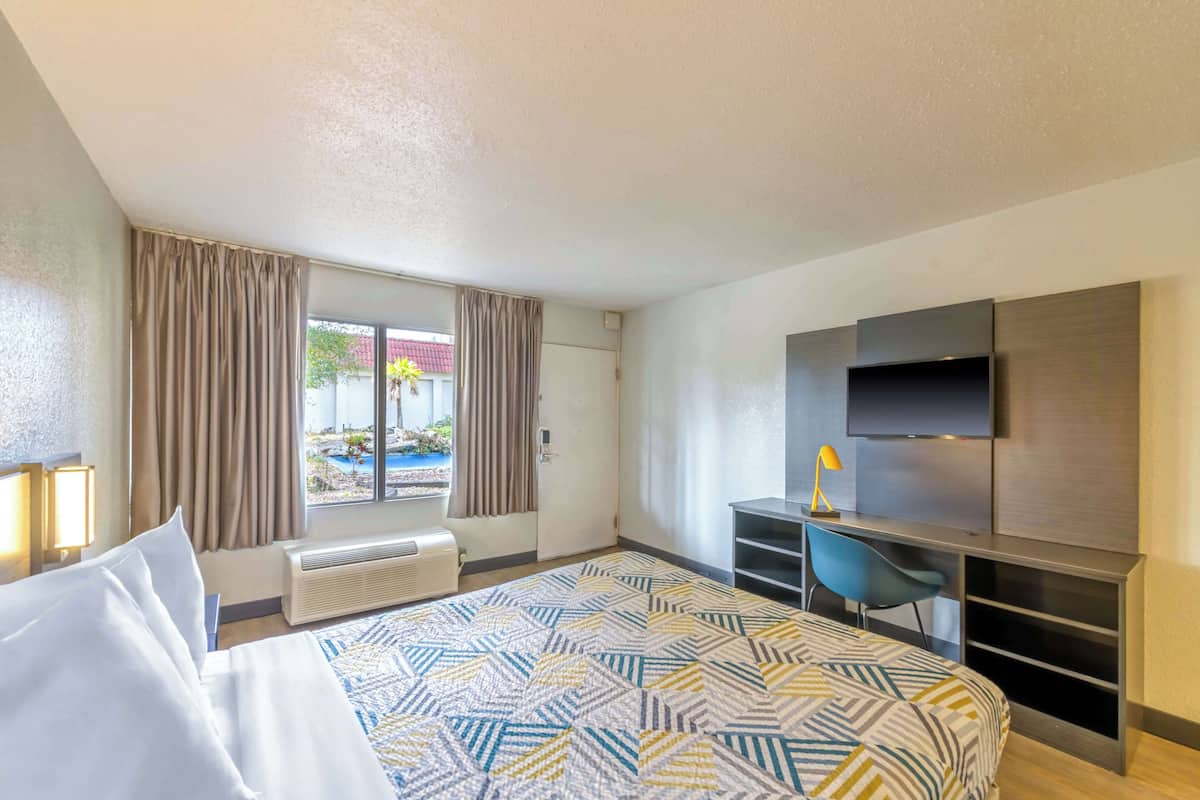 A hotel room with a bed, a desk, a chair, a TV, and a view of a pool and greenery.