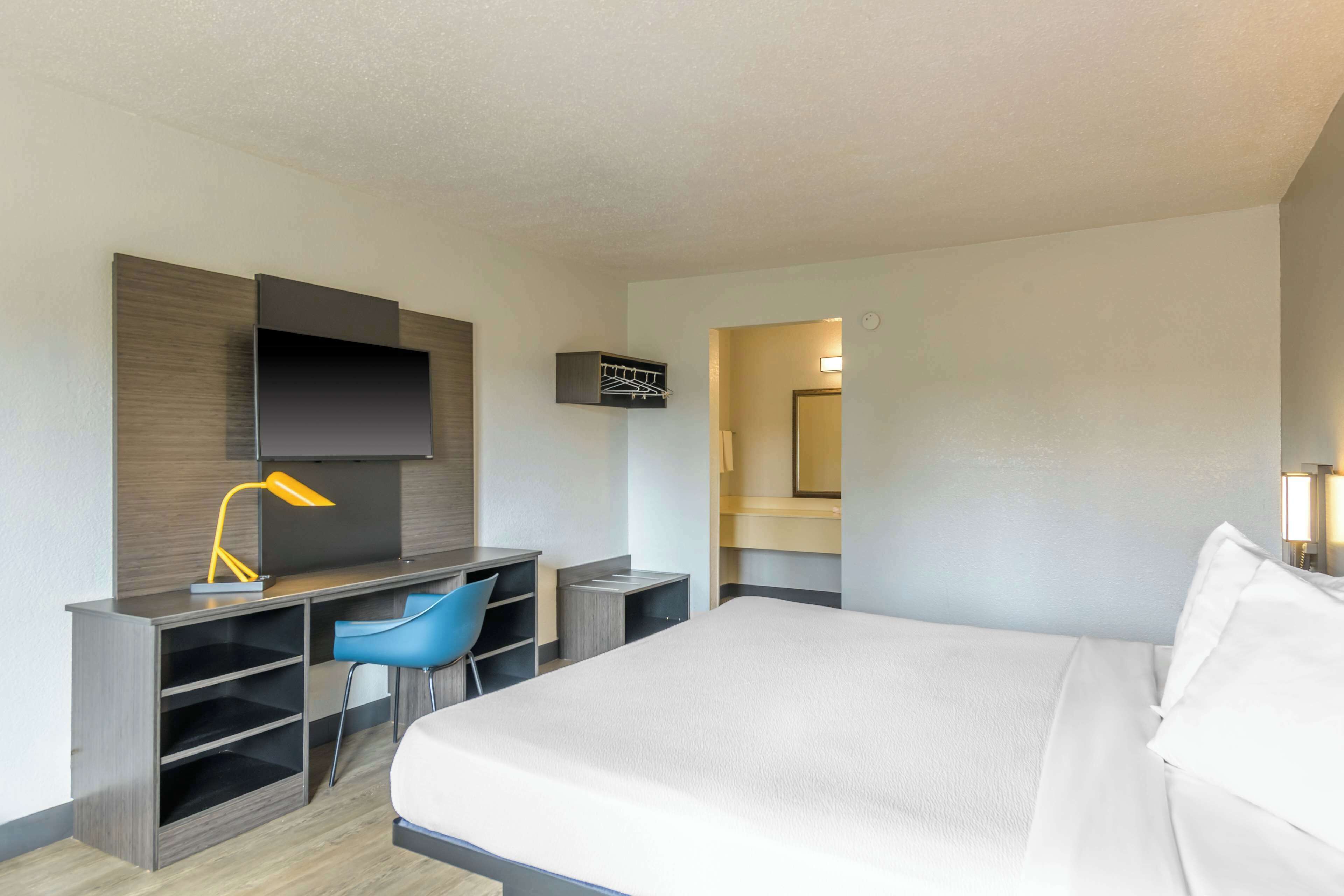 Ein Hotelzimmer mit einem Bett, einem Schreibtisch mit Stuhl, einem an der Wand befestigten Fernseher und einem kleinen Regal.