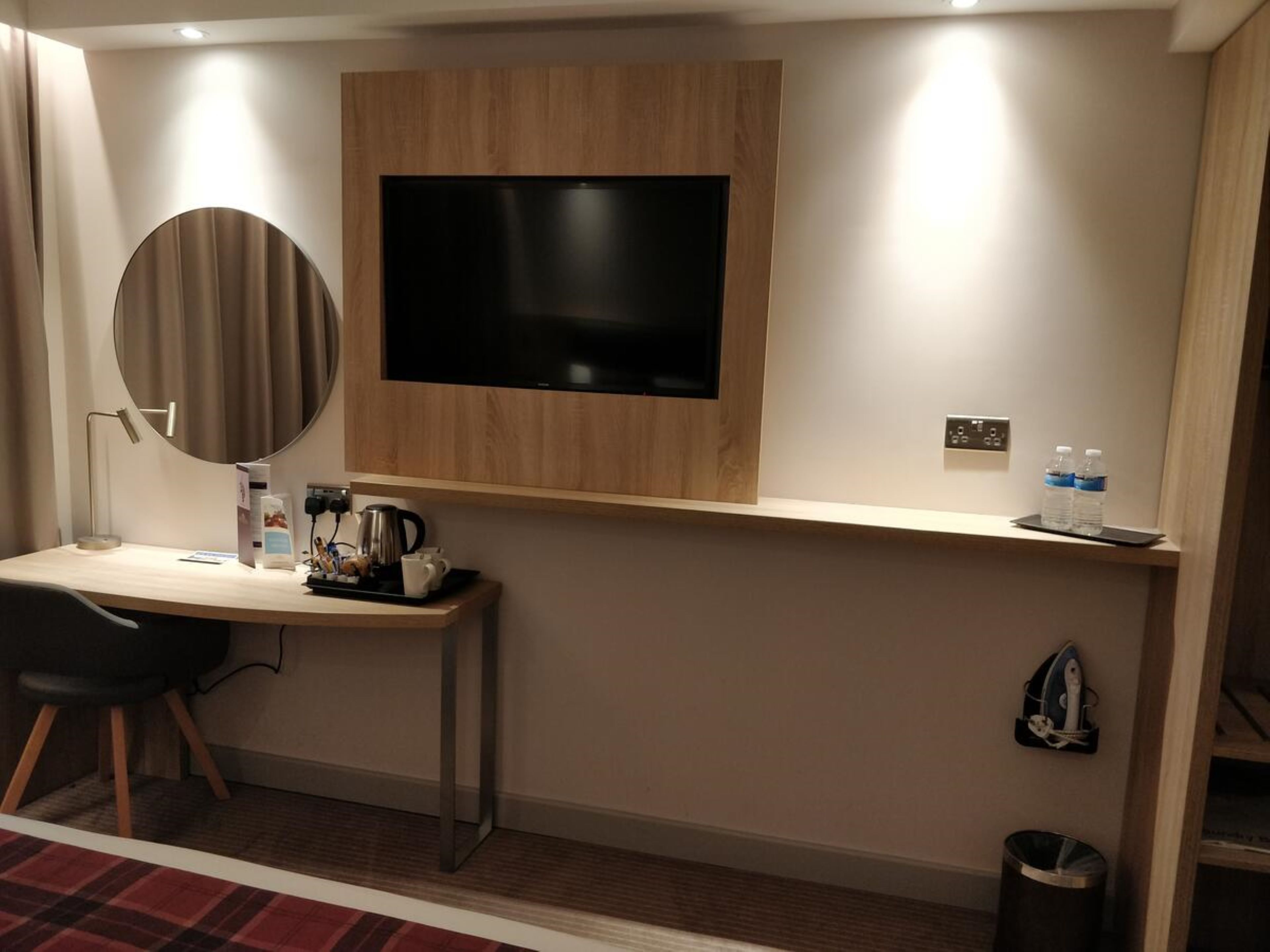 A hotel room with a flat-screen TV mounted on a wooden panel, a desk with a chair, a coffee maker, a kettle, a mirror, a trash bin, and a shelf with water bottles.