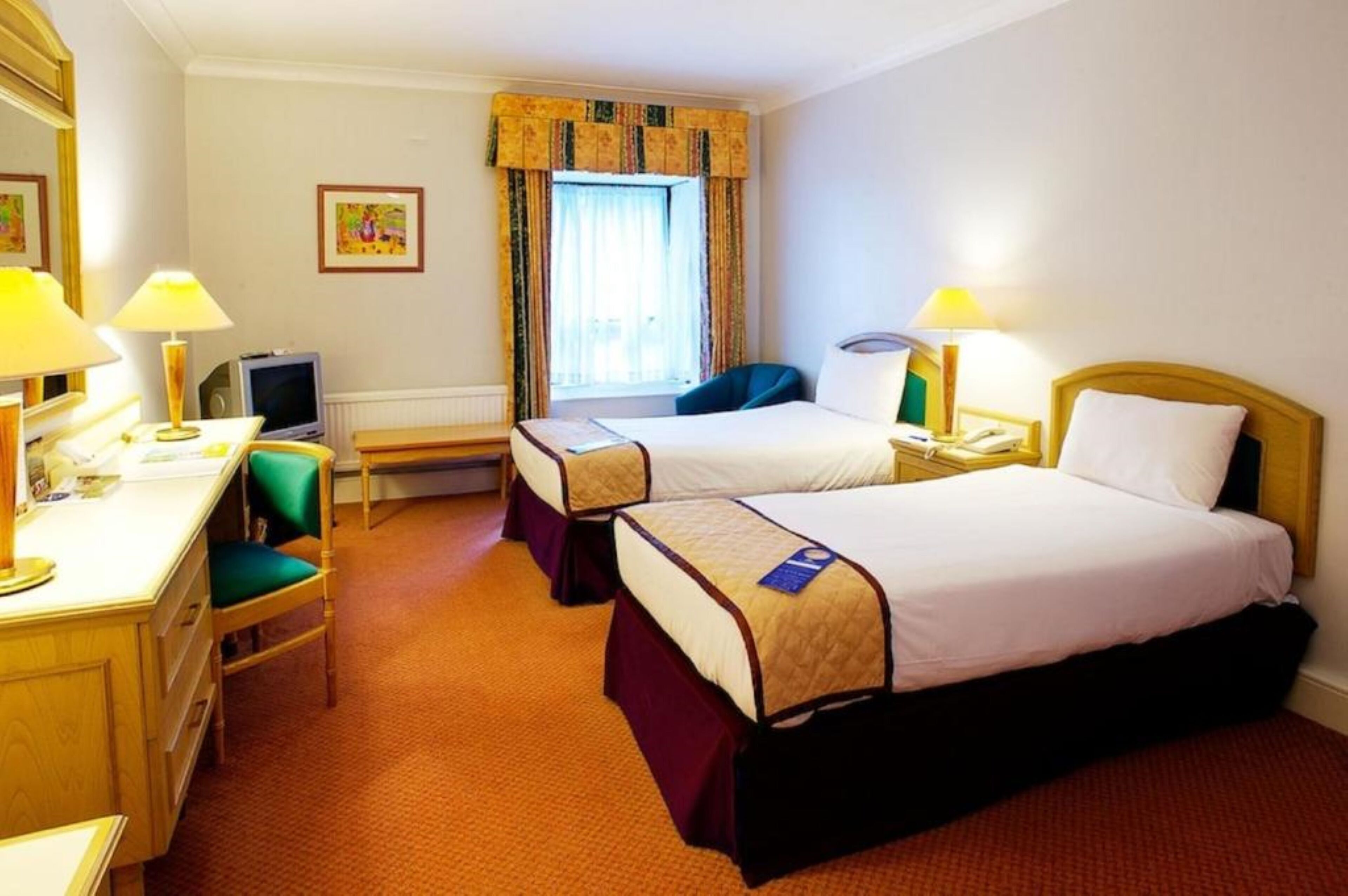A hotel room with two beds, a desk, a chair, a television, and a window with curtains.
