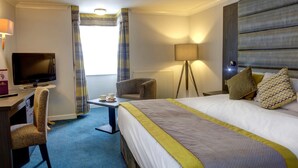 Una habitación de hotel con cama, escritorio con televisor, silla, lámpara y ventana con cortinas.