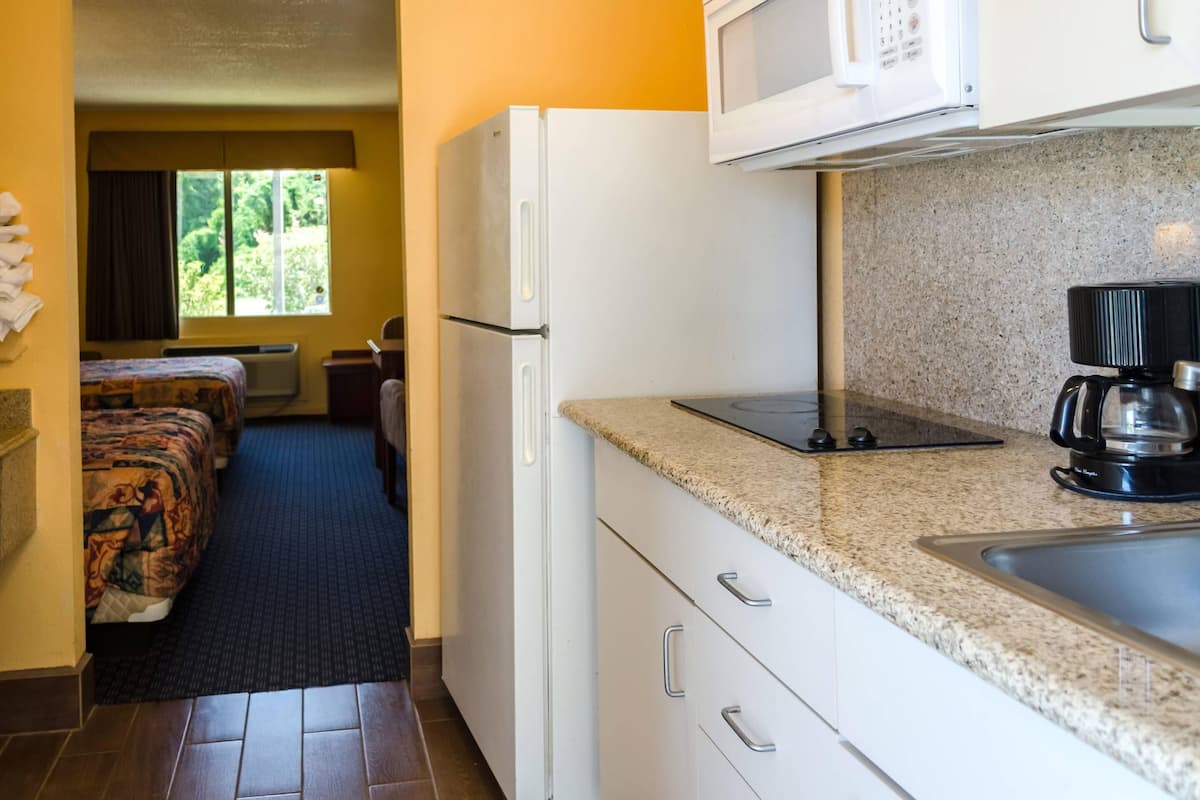 A compact hotel room with a kitchenette, a bed with a patterned bedspread, a window with curtains, and a view of greenery outside.
