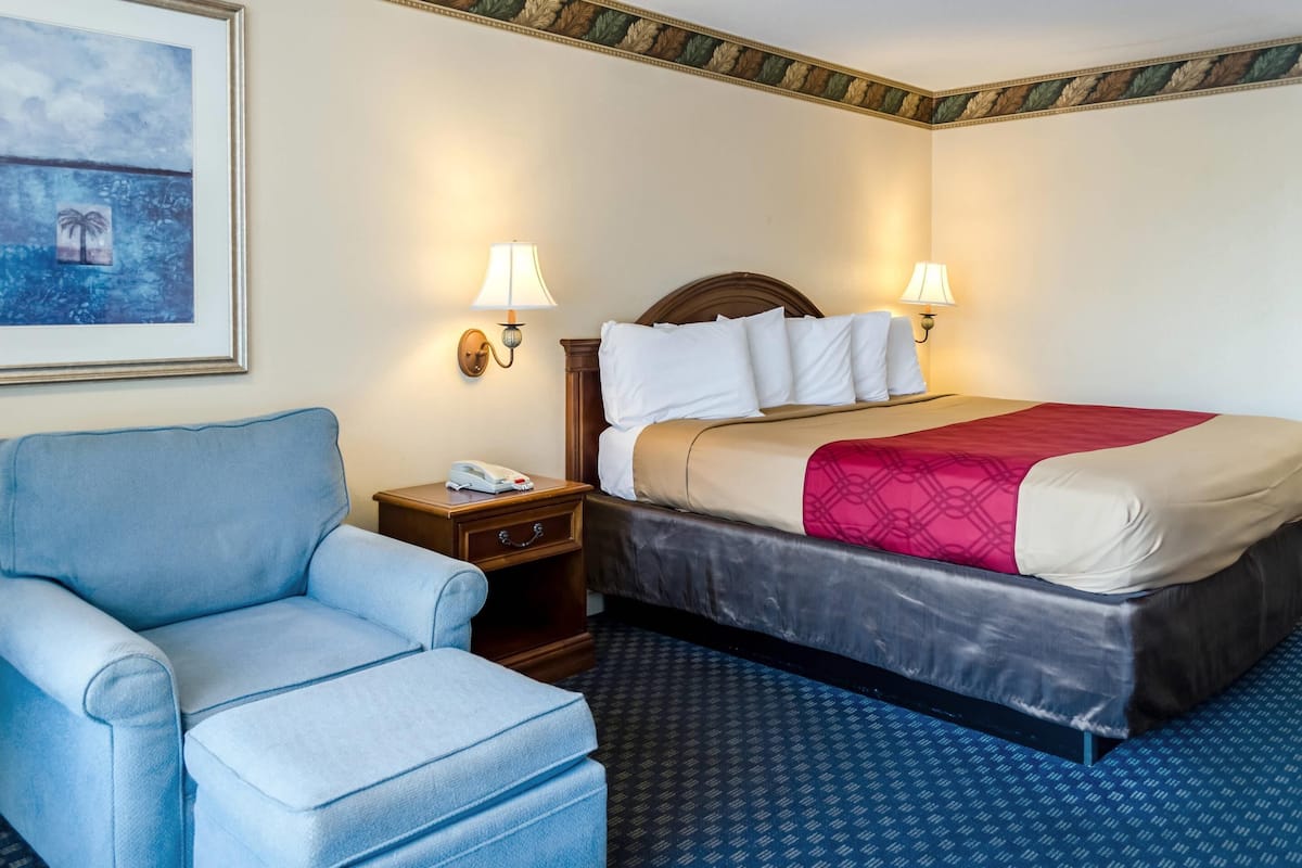 A hotel room with a bed, a blue armchair, a nightstand with a phone, and wall-mounted lamps.