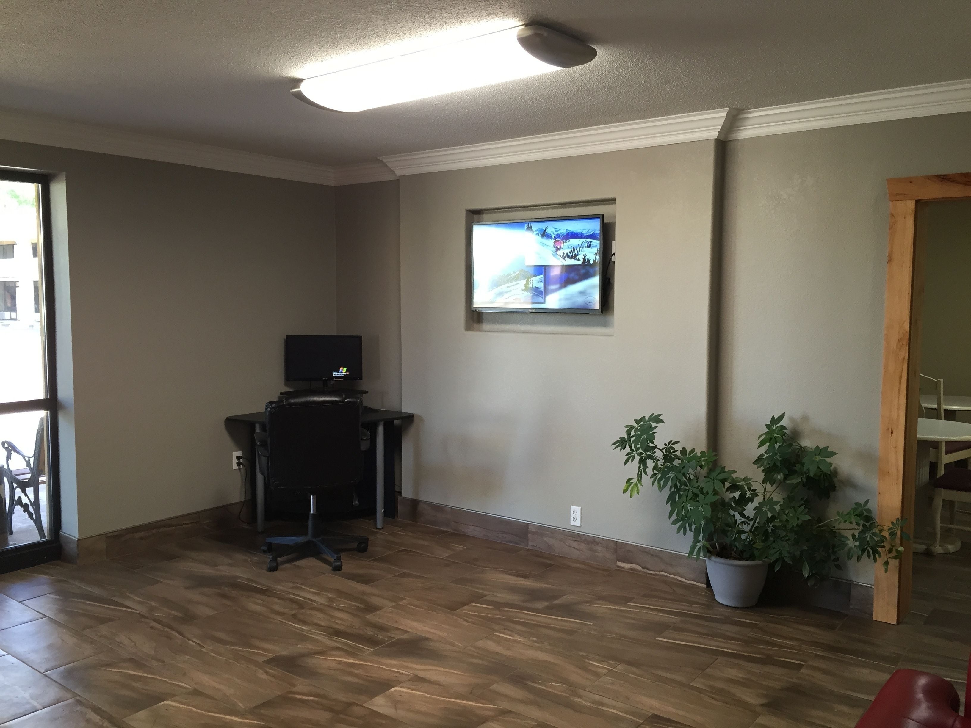 A room with a wooden floor, a desk with a computer, a potted plant, and a television mounted on the wall.